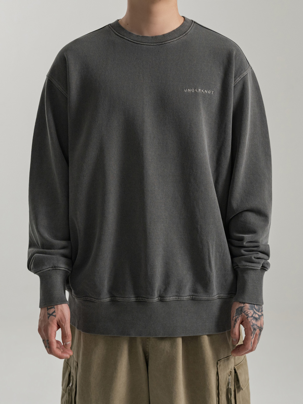 Real studio photograph of the UNDERKNOT wordmark-only sweatshirt in FADED GRAPHITE, worn by a model with the face out of frame