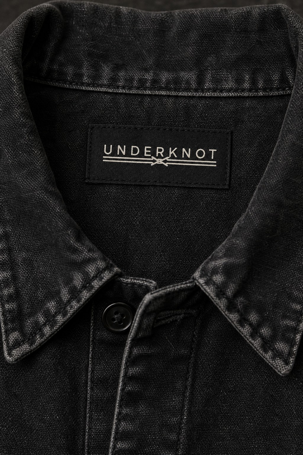 WASHED CHORE JACKET 넥 안쪽 중앙의 블랙 직조 UNDERKNOT 메인라벨과 웜 에크루 엠블럼을 보여 주는 디지털 디테일. 사이즈 표기 없음