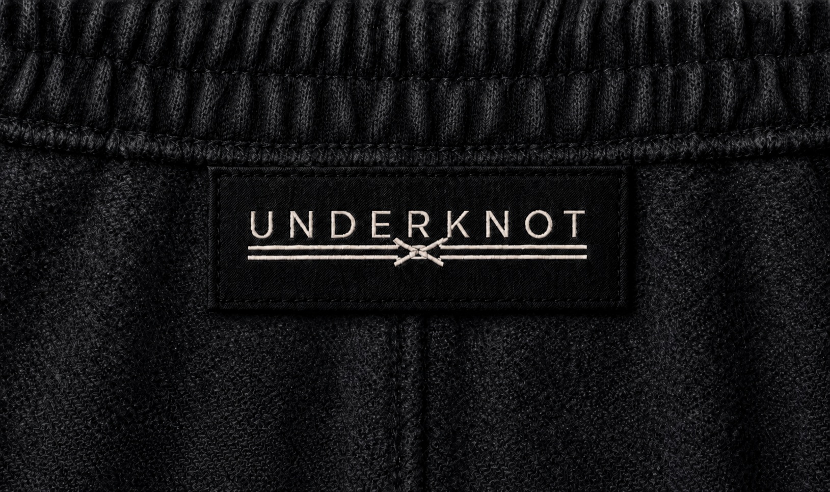 WIDE SWEATPANTS 뒤 허리 고무단 하단 봉제선에 연결된 블랙 직조 UNDERKNOT 메인라벨과 웜 에크루 엠블럼을 보여 주는 디지털 디테일. 사이즈 표기 없음